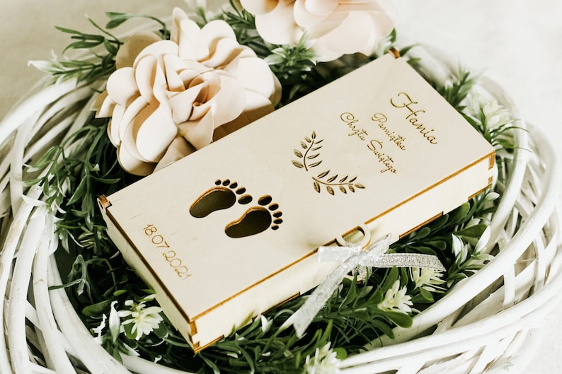 Puede incluir: Una caja de madera con fondo blanco y una cinta plateada. La caja est&aacute; grabada con el texto "Hania Pamiątka Chrztu Świętego" y la fecha "18.07.2021". La caja tambi&eacute;n presenta dos peque&ntilde;as huellas de pies.