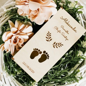 Puede incluir: Un rollo de madera con el texto "Aleksander Pamiątka Chrztu Świętego" y la fecha "22.08.2021". El rollo est&aacute; decorado con dos peque&ntilde;as ramas frondosas y un par de huellas de pies de beb&eacute;. El rollo est&aacute; rodeado de una cesta de mimbre blanca con hojas verdes y flores de color melocot&oacute;n.