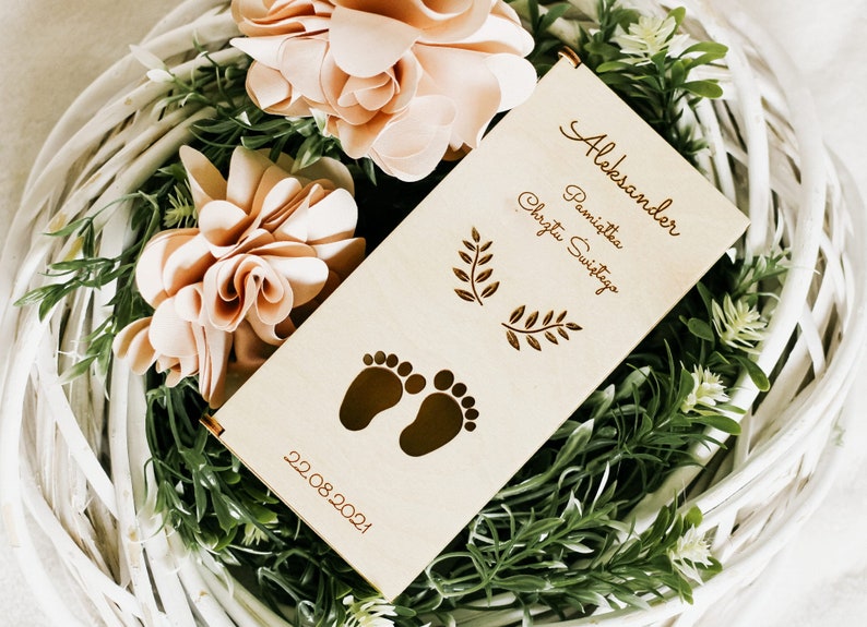 Puede incluir: Un rollo de madera con el texto "Aleksander Pamiątka Chrztu Świętego" y la fecha "22.08.2021". El rollo est&aacute; decorado con dos peque&ntilde;as ramas frondosas y un par de huellas de pies de beb&eacute;. El rollo est&aacute; rodeado de una cesta de mimbre blanca con hojas verdes y flores de color melocot&oacute;n.