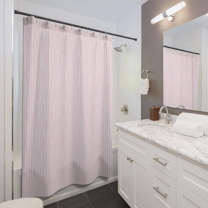 May include: A light pink and white striped shower curtain hangs in a bathroom. The curtain is suspended from a dark metal rod. The bathroom has white cabinets, a marble countertop, and a mirror.