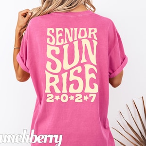 Puede incluir: Una camiseta de manga corta de color rosa brillante con un texto grande, ondulado y de color crema en la espalda que dice "SENIOR SUN RISE" con el a&ntilde;o "2*0*2*7" impreso debajo en la misma fuente y color, con la palabra "Crunchberry" en la esquina.