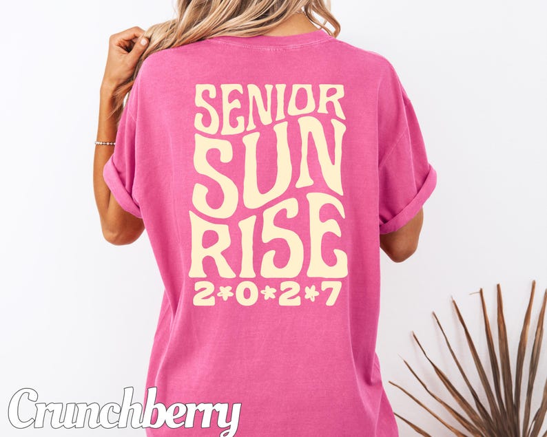 Puede incluir: Una camiseta de manga corta de color rosa brillante con un texto grande, ondulado y de color crema en la espalda que dice "SENIOR SUN RISE" con el a&ntilde;o "2*0*2*7" impreso debajo en la misma fuente y color, con la palabra "Crunchberry" en la esquina.