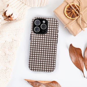 May include: A brown and white checkered phone case. The case is lying on a white surface, next to a wrapped gift, dried orange slice, and autumn leaves. A cream-colored sweater and cotton boll are also in the image.
