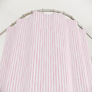 May include: A pink and white striped shower curtain hangs from a curved metal rod. The vertical stripes are hand-painted in a watercolor style, creating a soft, textured look. The curtain is made of a lightweight fabric.