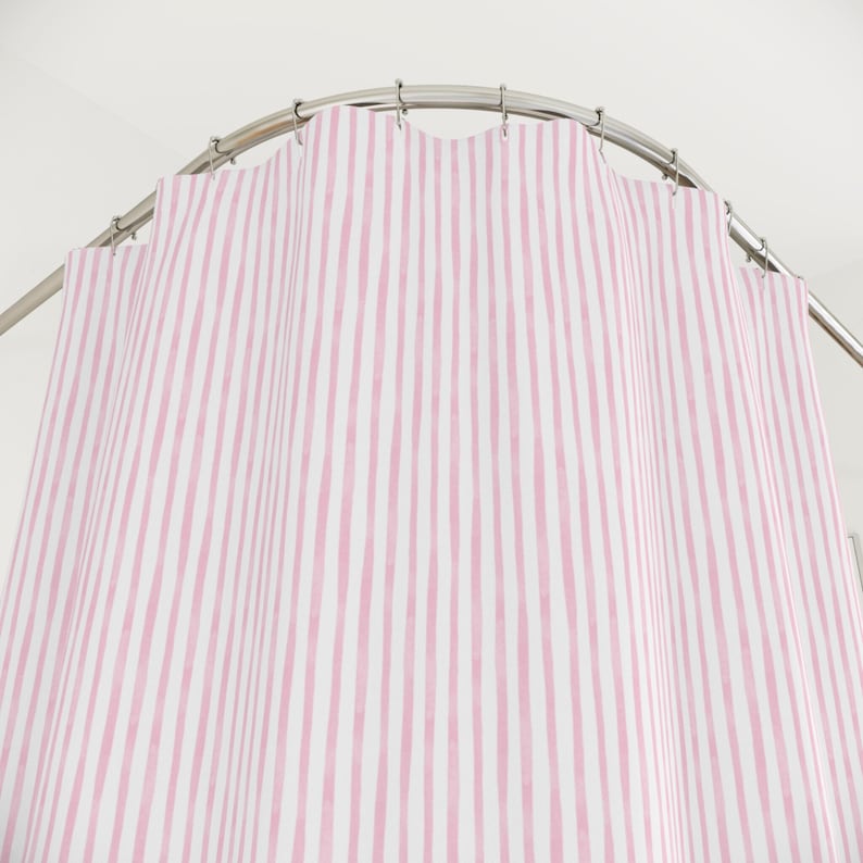May include: A pink and white striped shower curtain hangs from a curved metal rod. The vertical stripes are hand-painted in a watercolor style, creating a soft, textured look. The curtain is made of a lightweight fabric.