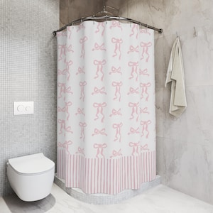 May include: A white shower curtain with a repeating pattern of pink bows. The bottom border features pink and white vertical stripes. The curtain is hanging in a bathroom with a white toilet and a gray tiled wall.