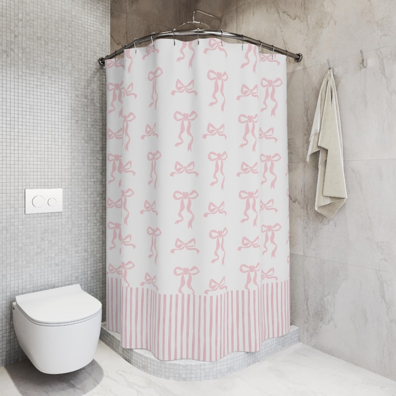 May include: A white shower curtain with a repeating pattern of pink bows. The bottom border features pink and white vertical stripes. The curtain is hanging in a bathroom with a white toilet and a gray tiled wall.