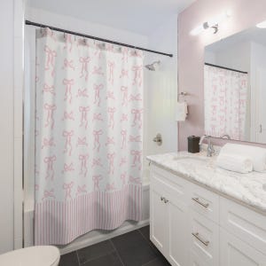May include: A white shower curtain with a repeating pattern of pink bows. The bottom of the curtain features pink and white vertical stripes. The bathroom has a white vanity, a mirror, and a shower head.