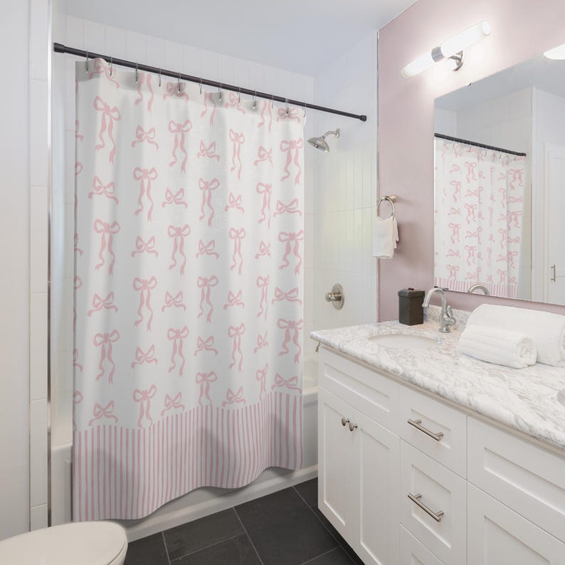 May include: A white shower curtain with a repeating pattern of pink bows. The bottom of the curtain features pink and white vertical stripes. The bathroom has a white vanity, a mirror, and a shower head.