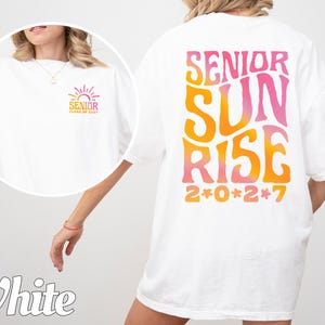 Puede incluir: Una camiseta blanca con un gr&aacute;fico grande en la espalda con el texto SENIOR SUNRISE 2*0*2*7 en una fuente ondulada con degradado de naranja a rosa. Una versi&oacute;n m&aacute;s peque&ntilde;a del mismo gr&aacute;fico est&aacute; impresa en la parte frontal izquierda.