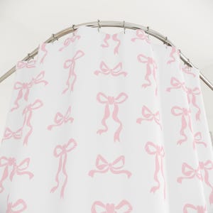May include: White shower curtain with a repeating pattern of pink bows. The bows vary in style and size, creating a playful and feminine design. The curtain hangs from a curved, silver-colored shower rod.