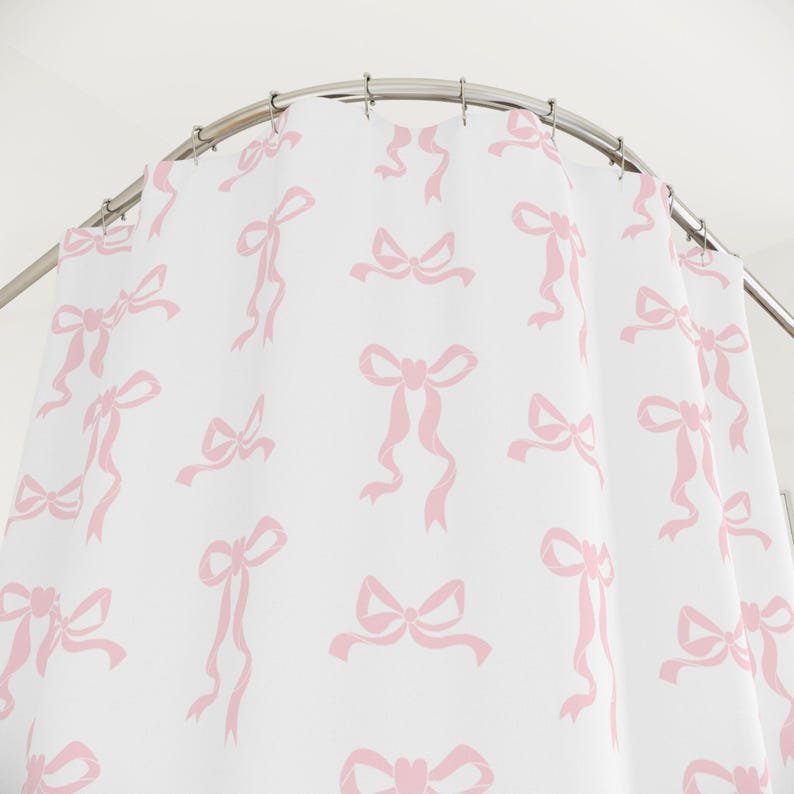 May include: White shower curtain with a repeating pattern of pink bows. The bows vary in style and size, creating a playful and feminine design. The curtain hangs from a curved, silver-colored shower rod.
