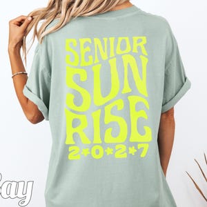 Puede incluir: Una camiseta de manga corta color verde salvia con texto amarillo ne&oacute;n brillante en la espalda que dice SENIOR SUNRISE 2*0*2*7 en una fuente ondulada y negrita. La camiseta tiene cuello redondo y mangas remangadas.