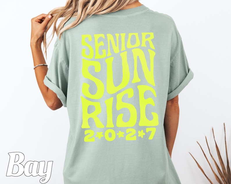 Puede incluir: Una camiseta de manga corta color verde salvia con texto amarillo ne&oacute;n brillante en la espalda que dice SENIOR SUNRISE 2*0*2*7 en una fuente ondulada y negrita. La camiseta tiene cuello redondo y mangas remangadas.