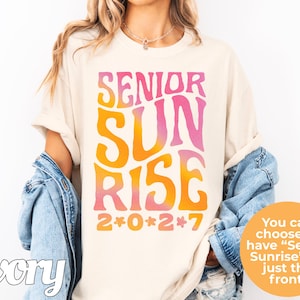 Puede incluir: Una camiseta de color marfil con el texto SENIOR SUNRISE en una fuente ondulada con degradado de rosa a naranja. Debajo del texto, el a&ntilde;o 2*0*2*7 est&aacute; impreso en naranja con peque&ntilde;os iconos de flores rosas separando cada d&iacute;gito.