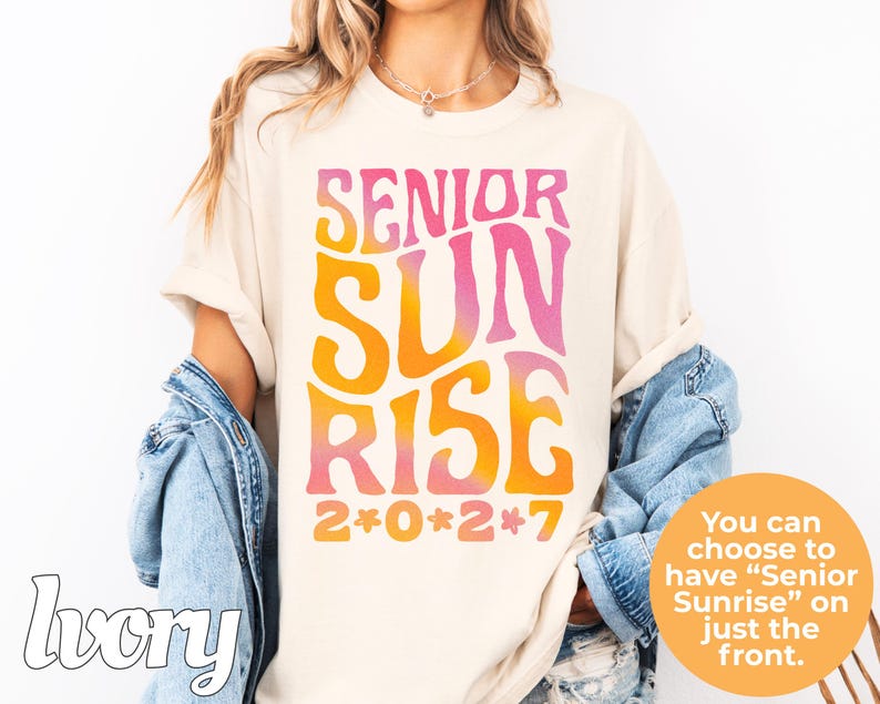 Puede incluir: Una camiseta de color marfil con el texto SENIOR SUNRISE en una fuente ondulada con degradado de rosa a naranja. Debajo del texto, el a&ntilde;o 2*0*2*7 est&aacute; impreso en naranja con peque&ntilde;os iconos de flores rosas separando cada d&iacute;gito.