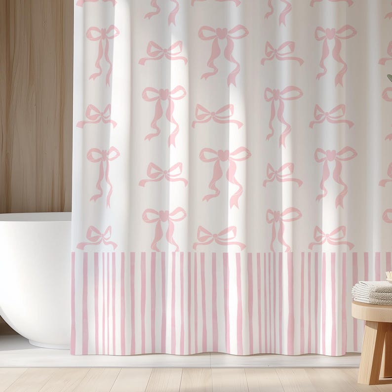 May include: White shower curtain with a repeating pattern of pink bows and ribbons. The bottom border features vertical pink and white stripes. The curtain is hanging in a bathroom setting, with a white bathtub visible.
