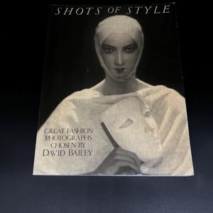Peut inclure: Couverture de livre avec le titre "SHOTS OF STYLE" et le texte "GREAT FASHION PHOTOGRAPHS CHOSEN BY DAVID BAILEY." La couverture présente une photographie en noir et blanc d'une personne portant un couvre-chef et tenant un masque.