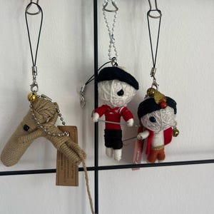 Puede incluir: Tres colgantes artesanales de hilo colgados de cordones negros. Uno es un caballo de cuerda beige con una campana, los otros son muñecos de hilo con trajes rojos y negros. Los muñecos tienen cintas negras en la cabeza y sostienen un alambre fino.