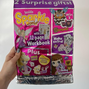 Op de afbeelding: Een roze en paarse Sparkle World-activiteitenverpakking met de tekst "2 Surprise gifts!" en "Inside Sparkle World". De verpakking bevat illustraties van een fee, poppen en een Furby-achtig wezen. Inclusief een werkboek van 12 pagina's en stickers.