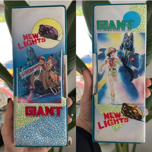 Puede incluir: Estuche vintage con borde turquesa. El estuche presenta ilustraciones coloridas de estilo anime y el texto "NEW LIGHTS" y "GIANT". La obra de arte representa personajes y un vehículo futurista.