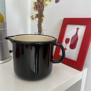 May include: A black enamel jug with a glossy finish, featuring a cream-coloured interior, a curved handle, and a small pouring spout. The jug has a cylindrical body and a smooth, reflective surface, showcasing a simple and functional design.
