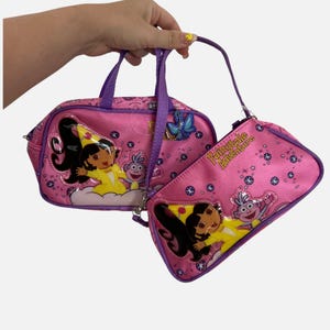 Op de afbeelding: Een roze handtas en een bijpassend kleiner tasje met Dora en Boots de aap. Beide items zijn versierd met een Fairytale Adventure-print, paarse randen en sterren. De handtas heeft paarse handvatten en een blauwe vlinderhanger als detail.