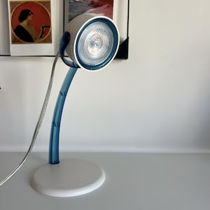 May include: A desk lamp featuring a white circular base and a curved, translucent blue neck. The lamp head is white with a deep blue rim surrounding a textured, reflective light bulb. A thin, silver-toned power cord extends from the lamp head.
