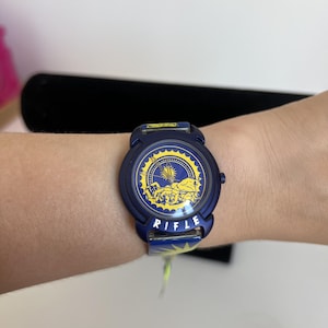 May include: A dark blue wristwatch featuring a circular face with a yellow sun and zodiac-style graphic design. The word RIFLE is printed in white capital letters on the lower part of the watch case. The watch band is also dark blue with yellow accents.