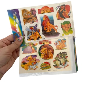 Puede incluir: Una colección de pegatinas vintage de El Rey León con Simba, Scar y otros personajes. Las pegatinas son coloridas y de varias formas, con el logotipo de El Rey León visible. Las pegatinas están en una funda de plástico transparente.
