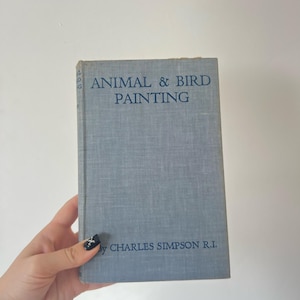 May include: A vintage book titled "ANIMAL & BIRD PAINTING" by Charles Simpson R.I. The book has a light blue cover with dark blue text. The book is held by a hand with a black and white nail design.