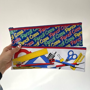 Pode incluir: Dois estojos. Um é azul com as palavras "Pencil Case" em um padrão repetido. O outro é branco com materiais escolares como uma régua, tesoura e apontador. Ambos têm uma borda vermelha.