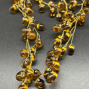 Op de afbeelding: Close-up van een decoratieve kralenketting. De ketting heeft amberkleurige, druppelvormige kralen met gele accenten, geregen op dun goudkleurig draad. De kralen zijn geclusterd, waardoor een textuurrijk ontwerp ontstaat.