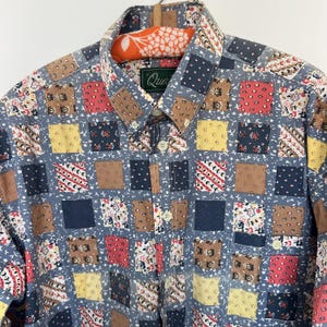 Op de afbeelding: Een overhemd met lange mouwen en knoopsluiting, voorzien van een veelkleurig patchworkpatroon van vierkanten in tinten blauw, rood, geel en bruin. Het overhemd heeft een puntige kraag, een knoopsluiting aan de voorzijde en een borstzak.