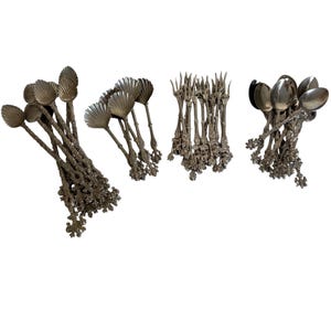 May include: A collection of silver-toned metal cutlery featuring ornate, textured handles with decorative ends. The set includes multiple spoons with shell-shaped bowls, several slender forks with pointed tines, and additional rounded spoons.