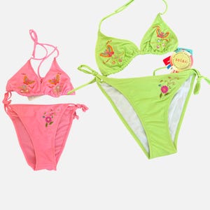 May include: Two two-piece bikini sets, one in bright pink and one in lime green. Each set features a triangle top with halter ties and a side-tie bottom, both decorated with embroidered butterfly and floral motifs. A circular tag reads solar.