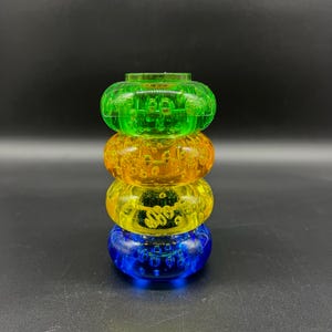 Op de afbeelding: Een stapel van vier transparante, kleurrijke plastic speelgoedringen. De ringen zijn groen, oranje, geel en blauw. Elke ring heeft een textuur met kleine, ronde inkepingen. De achtergrond is zwart.