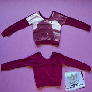 May include: Two maroon Adidas sweatshirts. One has a white and maroon graphic on the front. The other is solid maroon. The Adidas logo and "SAMPLE NOT FOR RESALE" are on a tag.