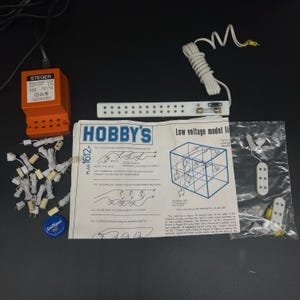 Op de afbeelding: Een set modeltreinbaanverlichtingcomponenten, waaronder een transformator, een lichtstrip en een set lampjes. De set bevat ook instructies voor het aansluiten van de componenten. De instructies bevatten een diagram van een modeltreinbaanlayout met de tekst "Hobby's Low Voltage Model Lighting" op de pagina.