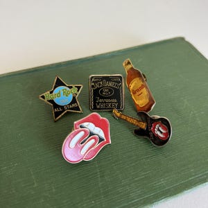 Może przedstawiać: Kolekcja pięciu metalowych przypinek emaliowanych, w tym czarna gwiazda z napisem Hard Rock Cafe, kwadratowy znak Jack Daniel's Tennessee Whiskey, butelka whisky Grant's, czerwono-białe logo języka oraz czarna gitara z logo języka.
