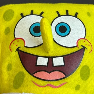 May include: Close-up of a yellow, square-shaped plush toy depicting a cartoon character's face. The character has large white eyes with blue irises, a long yellow nose, and a wide, smiling mouth with two square teeth. Red accents are around the eyes and mouth.