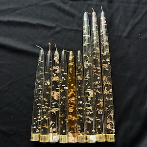 May include: A collection of clear, tapered candles with gold leaf and star-shaped accents. The candles have a transparent appearance, with gold foil and star-shaped glitter embedded within. The bases of the candles have white labels with black text.