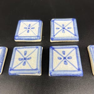 Puede incluir: Seis azulejos pequeños, cuadrados de cerámica con un diseño floral azul y blanco. Los azulejos tienen un fondo blanco con un patrón floral azul en el centro y un borde azul.