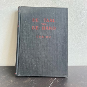 Puede incluir: Un libro antiguo titulado "DE TAAL VAN DE HAND" con el nombre del autor, CHEIRO, en letras rojas sobre una cubierta gris oscura. El libro está de pie sobre una superficie, mostrando su título.