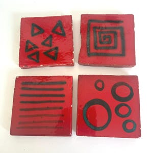 May include: Four red square coasters with black geometric designs. One coaster has triangles, another a spiral, a third has horizontal lines, and the last has circles. The coasters are likely made of ceramic or a similar material.