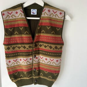 May include: A button-up, sleeveless sweater vest with a geometric pattern in olive green, red, tan, and cream. The vest is hanging on a white hanger. The pattern includes zigzags, diamonds, and horizontal stripes.