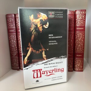Könnte beinhalten: Eine VHS-Kassette von The Royal Ballet's "Mayerling" mit Irek Mukhamedov und Viviana Durante. Das Cover zeigt eine Ballettaufführung in dunklen Farben mit goldenen Akzenten. Die Kassette befindet sich neben mehreren roten und goldenen Büchern.