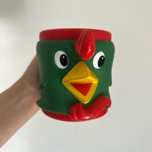 Puede incluir: Una taza con forma de pollo verde, rojo y amarillo. La taza presenta una cresta, un pico y una barbilla rojos, con grandes ojos blancos. La taza cilíndrica parece estar hecha de un material gomoso.