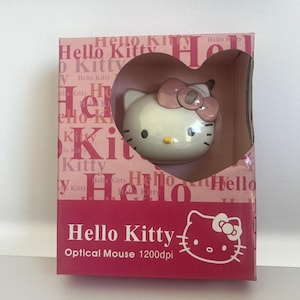 Puede incluir: Un ratón óptico Hello Kitty con sensor de 1200dpi, que presenta una cara blanca y un lazo rosa. El ratón está empaquetado en una caja de cartón rosa con un recorte en forma de corazón, mostrando el texto Hello Kitty Optical Mouse 1200dpi.
