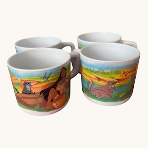 Puede incluir: Un juego de cuatro tazas de cerámica blanca con coloridas ilustraciones de la película Pocahontas. Los diseños incluyen al personaje Pocahontas, un mapache, un colibrí y nutrias, con el texto Pocahontas impreso en el lateral de las tazas.